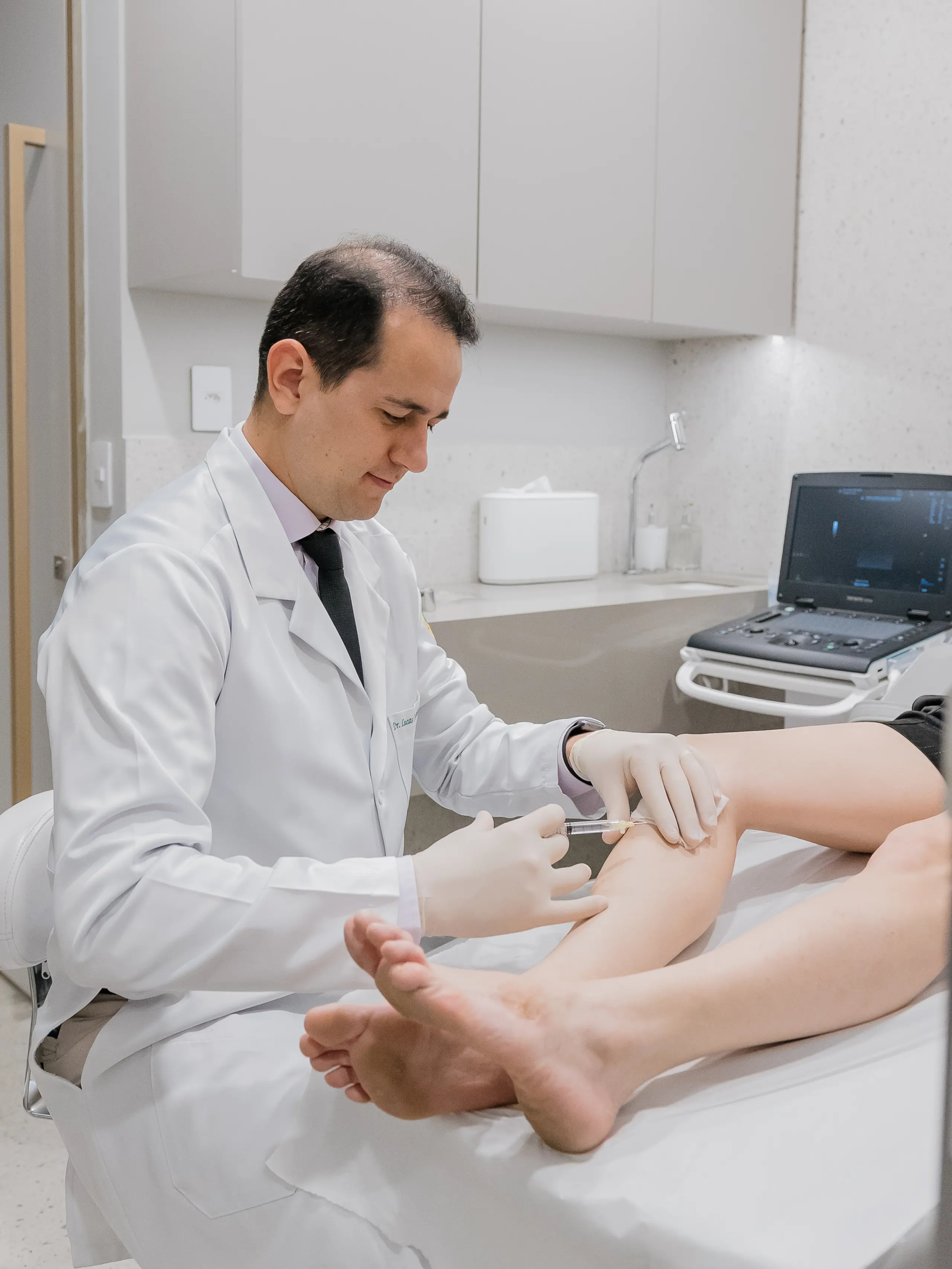 Dr. Lucas Cartafina aplicando escleroterapia na perna do paciente em seu consultório em Sorocaba